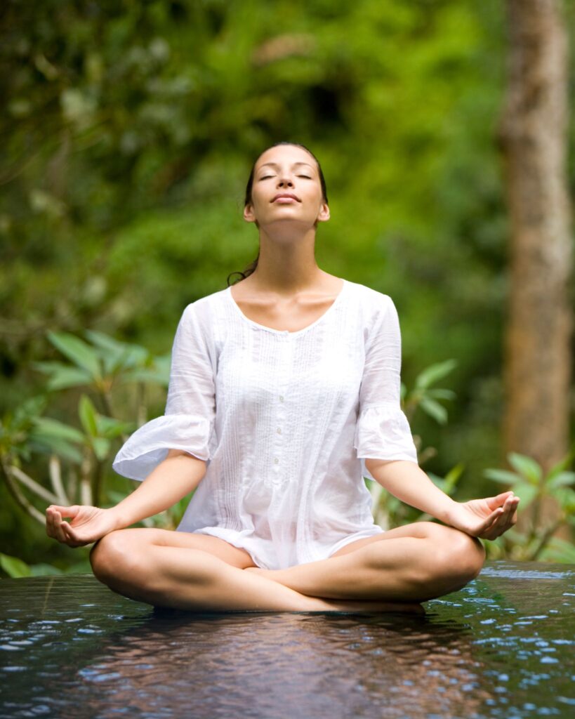 A woman sitting cross-legged in meditation outdoors, eyes closed, surrounded by greenery.